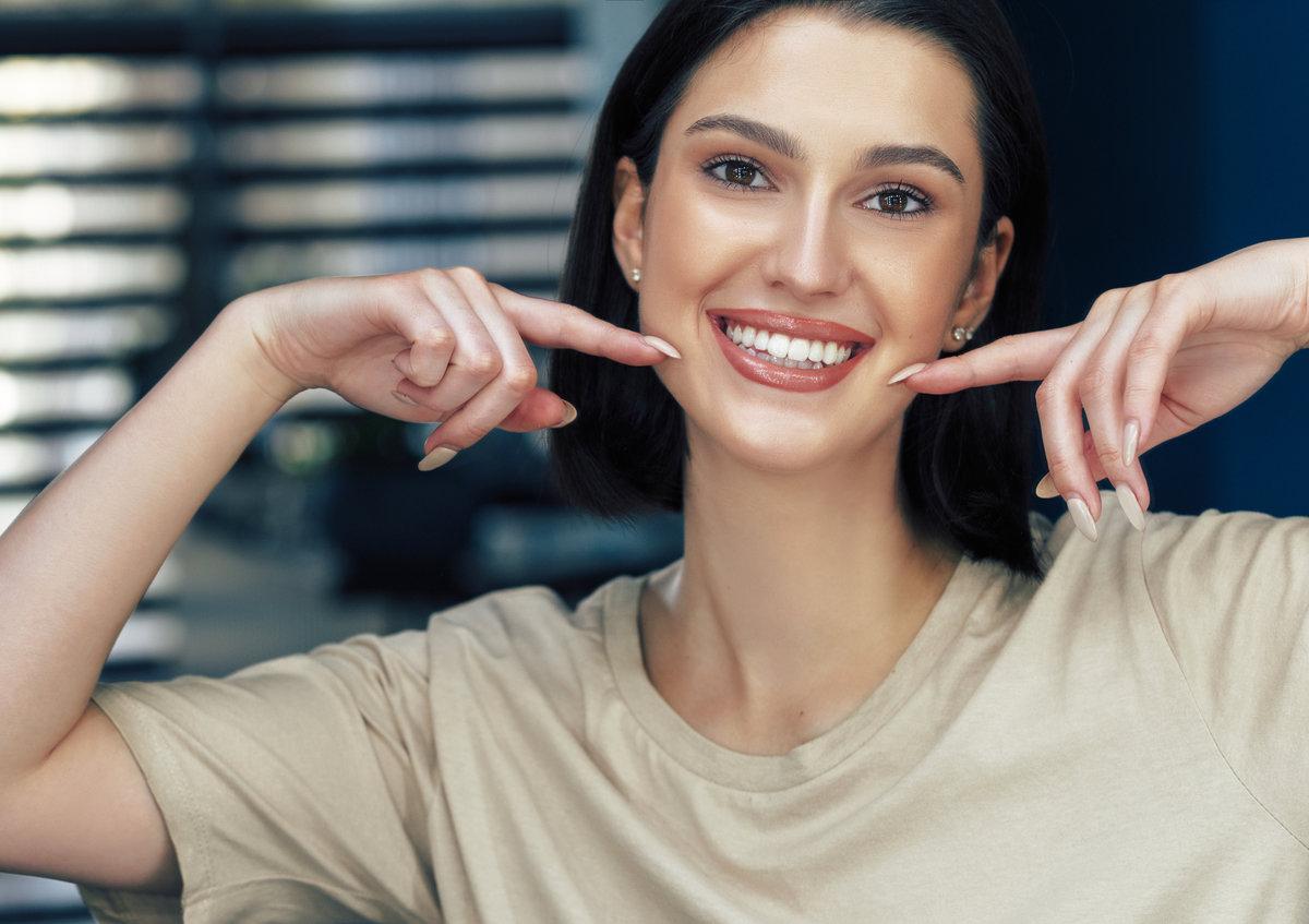 Portrait of a positive attractive girl with natural makeup smiling and showing with index fingers her healthy white teeth. Pretty young woman has joyful expression after makeup. Portrait of a positive attractive girl with natural makeup smiling and showing with index fingers her healthy white teeth. Pretty young woman has joyful expression after makeup.