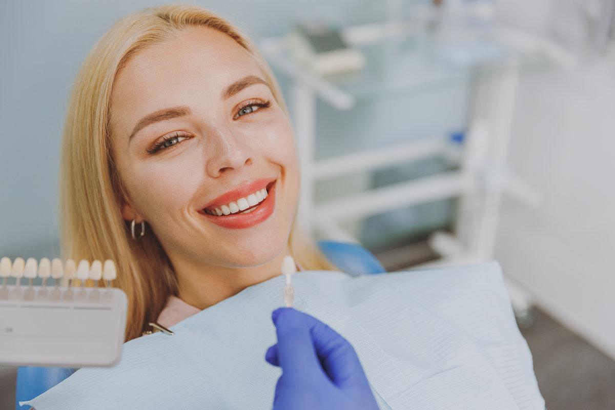 Close up young smiling woman choosing veneers enamel color palette with stomatologist compare teeth shade sit at dentist office chair indoor light modern cabinet Healthcare enamel whitening treatment. Close up young smiling woman choosing veneers enamel color palette with stomatologist compare teeth shade sit at dentist office chair indoor light modern cabinet Healthcare enamel whitening treatment