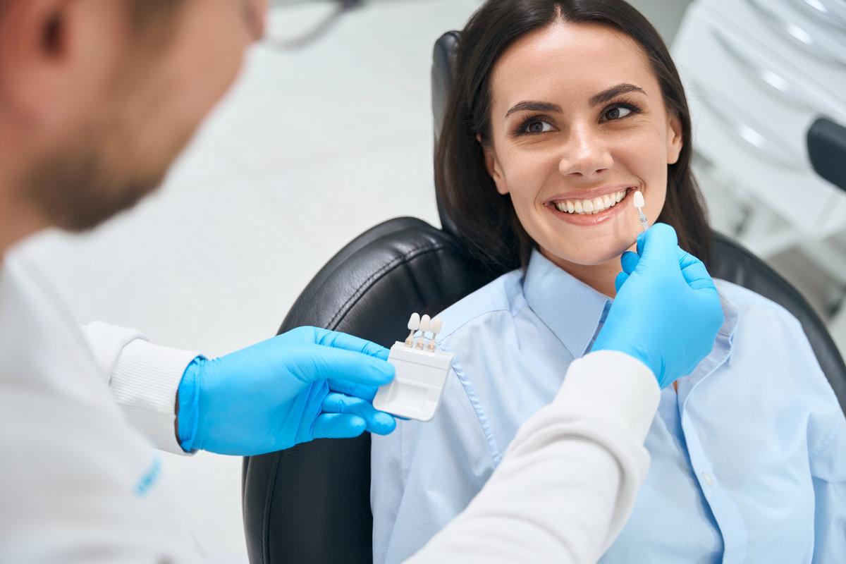 High-qualified dental technician choosing proper size and color of dental crown High-qualified dental technician choosing proper size and color of dental crown using some template, smiling woman client sitting in cozy orthodontic chair